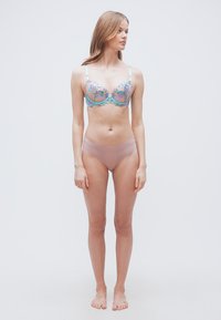 Γυναίκα που στέκεται ξυπόλυτη, φορώντας ένα σουτιέν με κεντήματα λουλουδιών σε ανοιχτό μπλε και ροζ χρώμα και ροζ εσώρουχα χωρίς ραφές, κοιτάζοντας προς τα πλάγια.