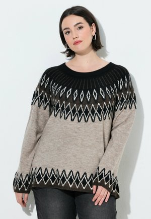 Kvinde med kort mørkt hår iført en beige sweater med sorte og hvide geometriske mønstre og mørke bukser, stående foran en hvid baggrund.