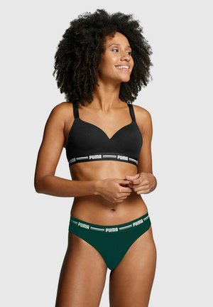 Smiling woman with curly hair wearing black Puma sports bra and dark green Puma underwear against a plain light background.