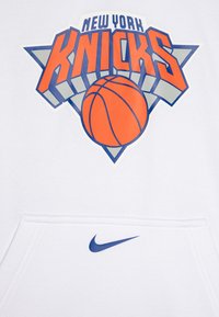 Bela kapuca z velikim modrim in oranžnim logotipom košarkarskega moštva New York Knicks, z majhnim modrim Nike swooshom pri žepu.
