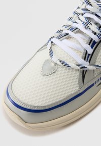 Baskets en mesh blanc avec accents argentés, rayures bleues et extérieur texturé. Elles comportent des lacets blancs et un détail de logo triangulaire argenté.