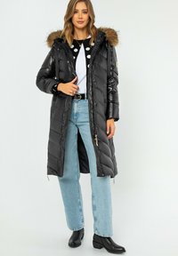 Schwarze Daunenjacke mit abnehmbarem Pelzkragen. Merkmale sind ein gestepptes Muster, vordere Reißverschlüsse und Seitentaschen, kombiniert mit hellblauen Jeans.