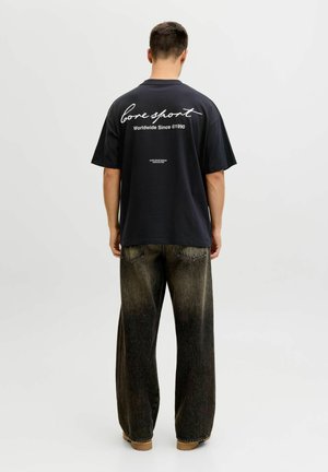 Mees seisab seljaga vaataja poole, kandes musta ülepõlve T-särki valge tekstiga ja lahtiseid pleekinud musti teksapükse, taust on ühevärviline.