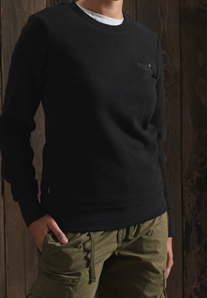 Osoba ubrana w czarną koszulę z długim rękawem na białej podkoszulce oraz oliwkowo-zielone spodnie bojówki, stojąca z rękami w kieszeniach na tle drewnianej ściany.