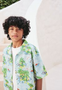 Chemise à manches courtes avec boutons, en tissu bleu clair orné d'un imprimé tropical de palmiers et de scènes aquatiques. Maillot blanc visible en dessous.