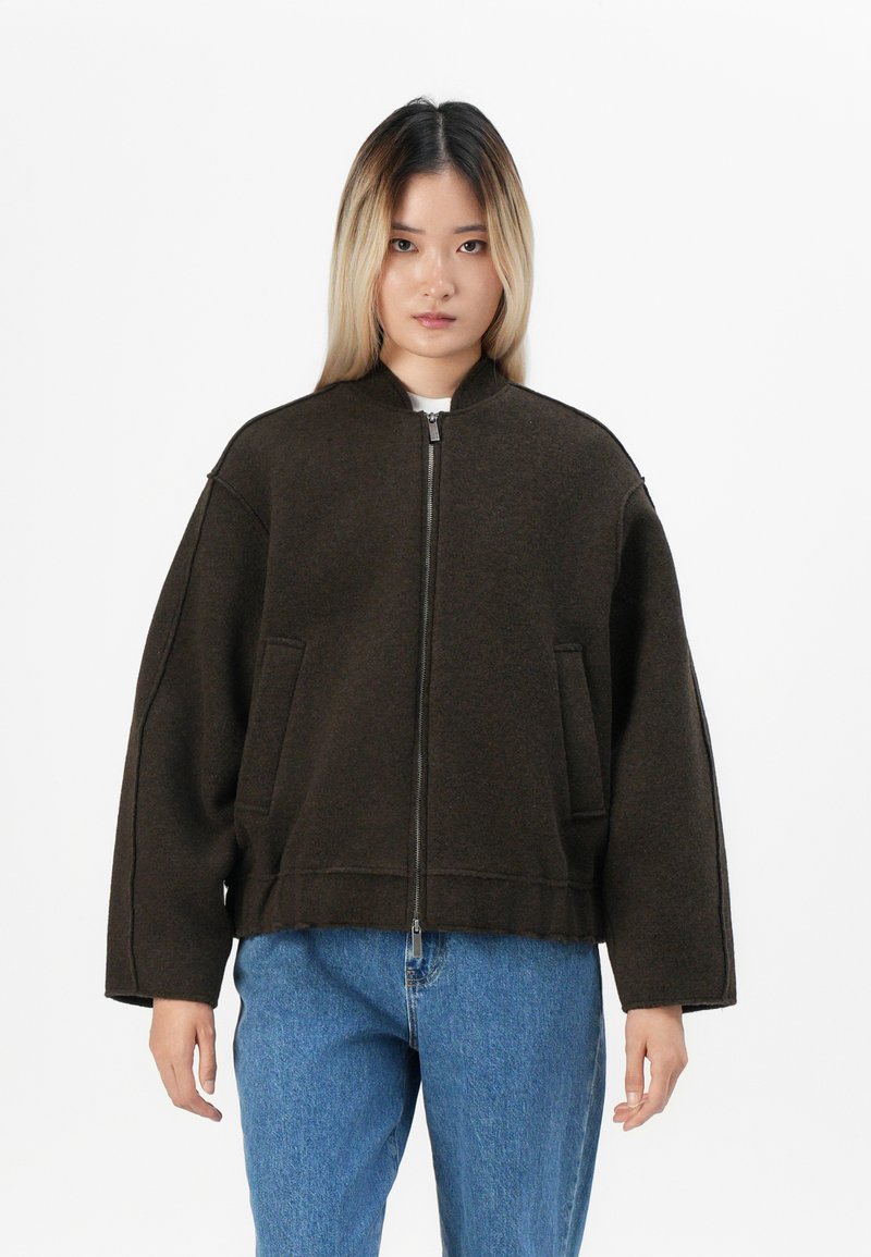 Brun zip-up jakke i et tykt stoff med høy hals, oversized passform, sidelommer og synlige sømdetaljer. Båret over blå jeans.
