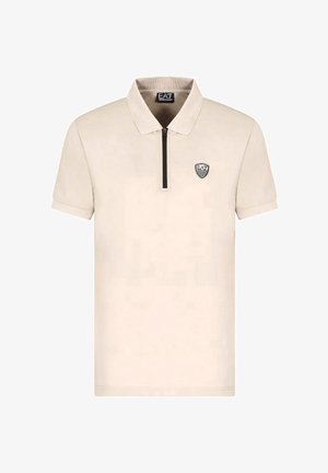 Polo shirt beige a maniche corte realizzato in tessuto morbido, presenta un colletto con zip e un patch logo nero sul lato sinistro del petto.