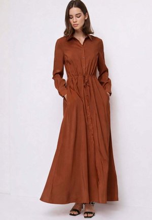 Donna che indossa un abito lungo color ruggine con bottoni, vita con coulisse e maniche lunghe, in piedi contro uno sfondo chiaro e semplice.