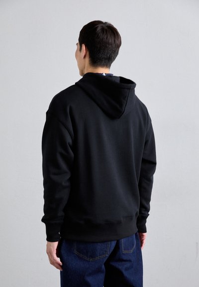 Schwarzer Kapuzenpullover in lässigem Schnitt, mit einer Kordelzugkapuze und gerippten Bündchen. Trägt ihn über blauen Jeans, was ein sauberes, minimalistisches Design zur Schau stellt.