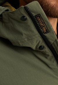Olijfgroene jas met een rits- en drukknoopsluiting, met een zwarte rits en een logopatch op de kraag. Glad materiaal met een matte afwerking.