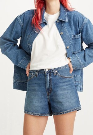 Person med rødt hår iført en hvid t-shirt, blå denimjakke og højtaljede blå denimshorts med hænderne i lommerne.