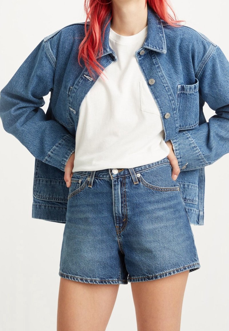 Personne aux cheveux roux portant un t-shirt blanc, une veste en jean bleue et un short en jean bleu taille haute avec les mains dans les poches.
