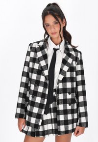 Černo-bílý kostkovaný blazer s dvouřad�ým zapínáním, doplněný bílou košilí a černou kravatou, vyrobený z texturované vlněné tkaniny.