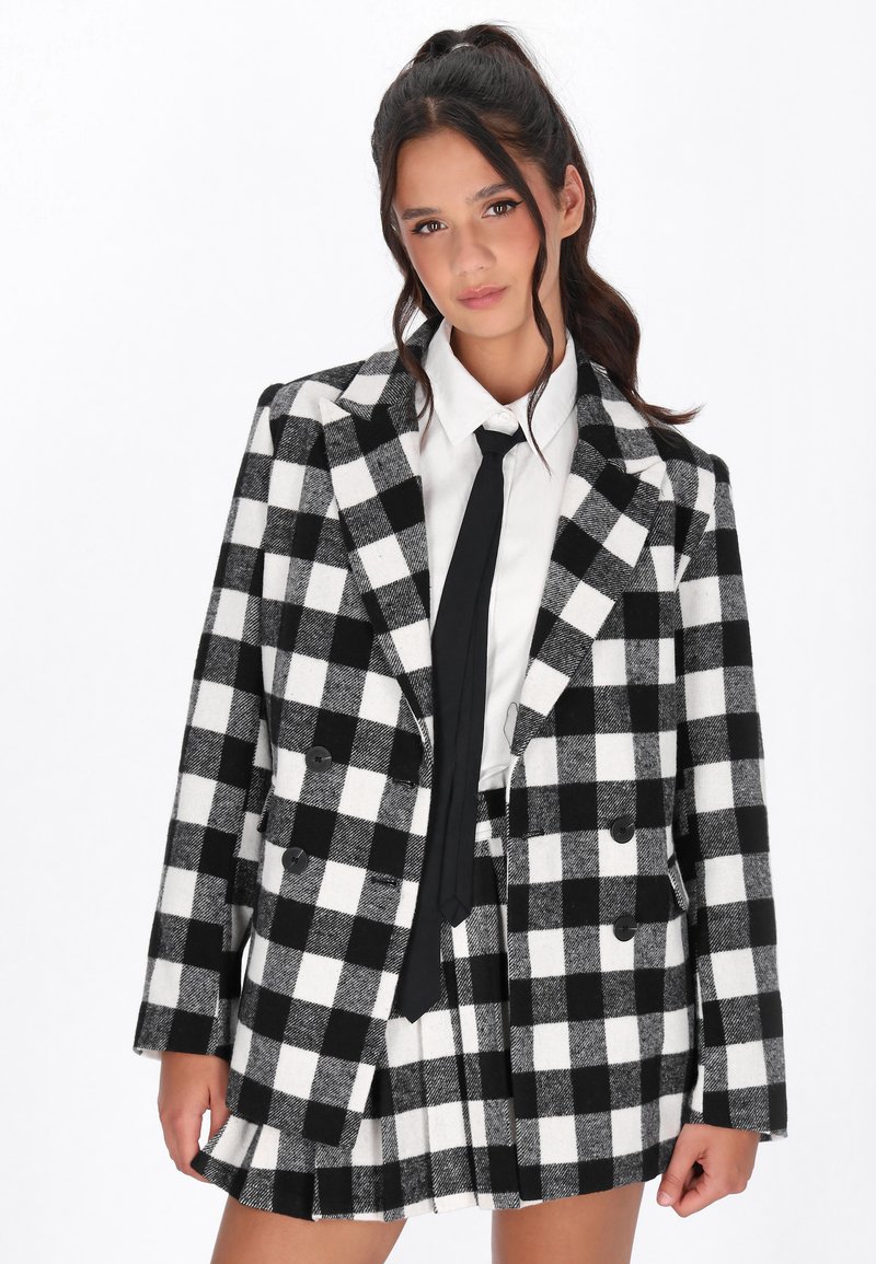 Černo-bílý kostkovaný blazer s dvouřadým zapínáním, doplněný bílou košilí a černou kravatou, vyrobený z texturované vlněné tkaniny.
