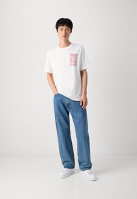 Hvid T-shirt med korte ærmer og et rød grafisk design på venstre bryst; sammen med blå løstsiddende jeans og hvide sneakers.