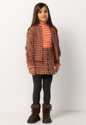 Meisje in een bruine geruite jas en rok, met een gestreepte perzikkleurige top, donkere geribde maillot en bruine laarzen met bontvoering, staand tegen een witte achtergrond.