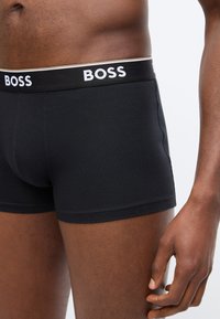 Buste masculin et haut des cuisses portant un boxer noir de la marque BOSS avec une ceinture noire arborant le lettrage blanc BOSS.