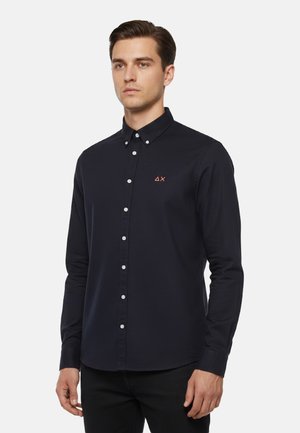 Uomo che indossa una camicia nera aderente con piccoli bottoni e un piccolo logo sul petto sinistro, in piedi davanti a uno sfondo chiaro uniforme.