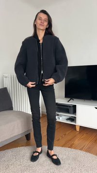 Casaco bomber de lã negra com fecho de correr frontal, mangas oversized e gola canelada, combinado com calças pretas justas e sapatos planos pretos.