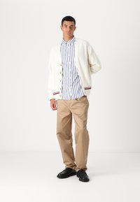Hvit strikket cardigan med gullknapper, lagdelt over en blå og hvit stripet skjorte. Beige bukser og svarte sko fullfører antrekket.