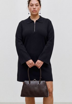 Femme portant une robe noire à manches longues avec col zippé, tenant un sac à main en cuir marron foncé par ses anses devant ses jambes.