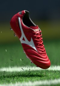 Crampons de soccer rouges en cuir, avec des accents blancs, une surface texturée et six pointes métalliques. De l'herbe et des débris sont visibles en dessous.