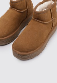 Botas de tobillo de ante marrón con un interior suave y esponjoso, punta redondeada y suela de goma texturizada y acanalada, con un diseño informal.