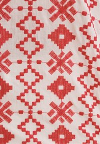 Vyšívaná textilie s červeným geometrickým vzorem na světle béžovém pozadí, která zobrazuje složité vzory a různé textury.