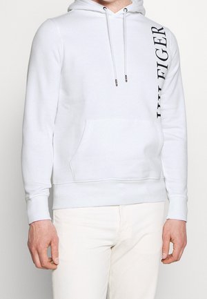 Hvid pullover-hoodie med forlommer og sort lodret "TOMMY HILFIGER" tekst på venstre skulder, båret med hvide bukser.