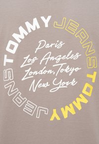Λογότυπο Tommy Jeans σε λευκούς και κίτρινους κύκλους με μια λίστα πόλεων: Παρίσι, Λος Άντζελες, Λονδίνο, Τόκιο, Νέα Υόρκη σε μπεζ υφασμάτινο φόντο.