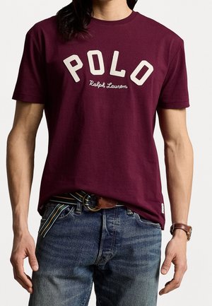 Homme portant un t-shirt Polo Ralph Lauren bordeaux à manches courtes, un jean bleu, une ceinture rayée et une montre marron.