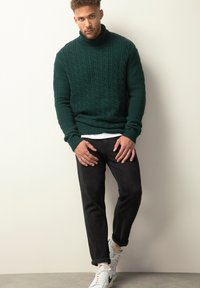 Dunkelgrüner Kabelstrickpullover mit hohem Kragen, kombiniert mit schwarzen schmal geschnittenen Jeans und weißen Turnschuhen mit grauen und beigen Akzenten.