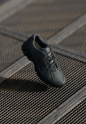 Zapatilla negra New Balance con suela texturizada, cordones atados, colocada sobre una superficie de rejilla metálica con líneas diagonales bajo iluminación suave.