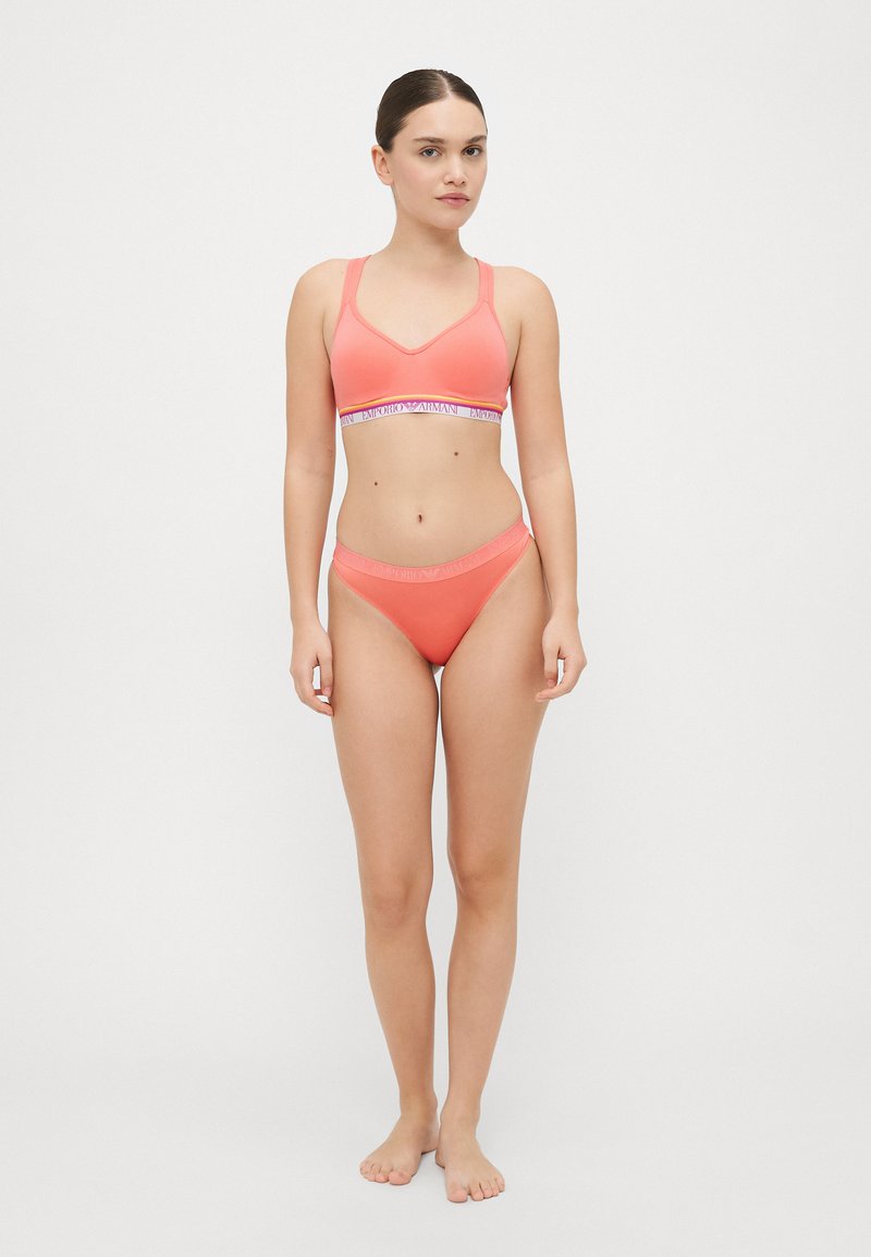 Coral-colored two-piece lingerie set featuring a v-neck bralette and matching hipster briefs, with an elastic waistband displaying the brand name.