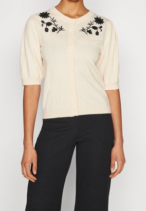 Femme portant un cardigan crème à manches courtes boutonné avec une broderie florale noire près des épaules et un pantalon noir.