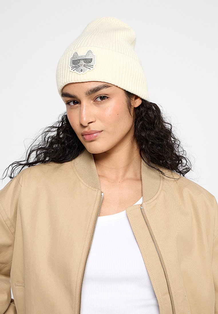 Frau mit lockigem, dunklem Haar trägt eine cremefarbene Mütze mit einem schwarz-weißen Katzen-Patch, eine beige Jacke und ein weißes Oberteil vor einem schlichten Hintergrund.