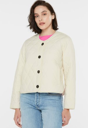 Woman wearing a quilted cream jacket with black buttons over a pink top and light blue jeans, standing against a plain white background.
