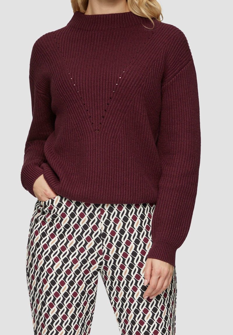Bordeaux gebreide trui met een hoge hals en V-vormige uitsparingen. Gepaard met gestreepte broek met geometrische vormen in zwart, wit en roze.