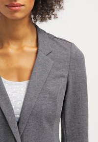 Blazer gris en tissu doux, avec un col à revers et une coupe ajustée, porté sur un haut gris clair à pois.