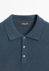 Tmavě modré pletené polo tričko s klasickým límcem, se zapínáním na knoflíky a čtyřmi stříbrnými knoflíky. Měkká textura, pečlivé zpracování.