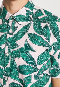 Camicia a maniche corte rosa chiaro con bottoni, caratterizzata da una grande stampa di foglie tropicali verdi e una tasca sul petto.