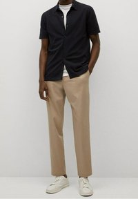 Lyhythihainen laivastonsininen paita, jossa napitus edessä, yhdistetty vaaleanruskeisiin housuihin ja valkoisiin lenkkareihin. Sileä kangas, rento istuvuus, hillitty yksityiskohdat.