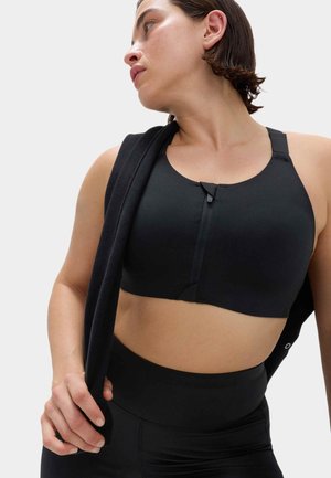 Kvinde i sort sport-bh med lynlås foran og leggings med høj talje, holder en sort jakke over skulderen og kigger til siden.