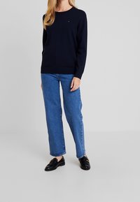 Tmavě modrý svetr s dlouhým rukávem, kulatým výstřihem a žebrovaným lemem, doplněný o světle modré džíny s vysokým pasem a rovnými nohavicemi, a černé mokasíny.