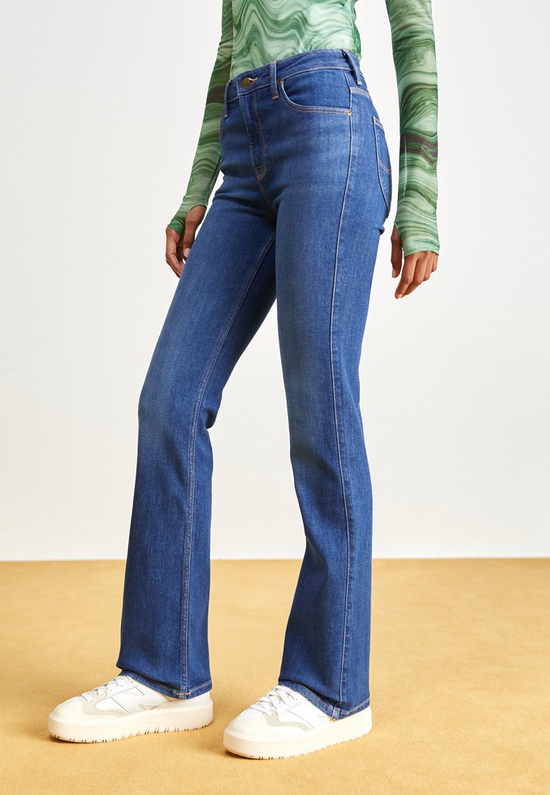 Pantalones vaqueros acampanados de mezclilla en azul con cintura alta. Cuenta con cinco bolsillos, costuras doradas y un ligero estiramiento. Combinados con zapatillas blancas.
