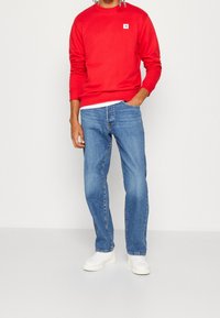 Sudadera roja con un pequeño logo en el pecho, combinada con vaqueros azules de corte recto y zapatillas blancas, posando frente a un fondo claro.