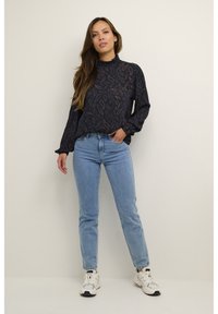 Marinebluse med et tekstureret mønster, høj hals og lange ærmer, parret med lyseblå jeans og hvide sportssko.