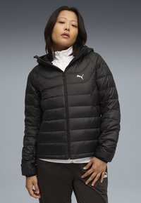 Giacca a vento nera con zip frontale e cappuccio, caratterizzata da un design trapuntato e un logo bianco sul lato sinistro del petto. Texture liscia e lucida.