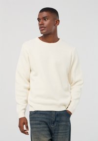 Maglione a maglia colore crema con colletto tondo, maniche lunghe e polsini a coste; indossato con jeans in denim blu; texture liscia, design semplice.