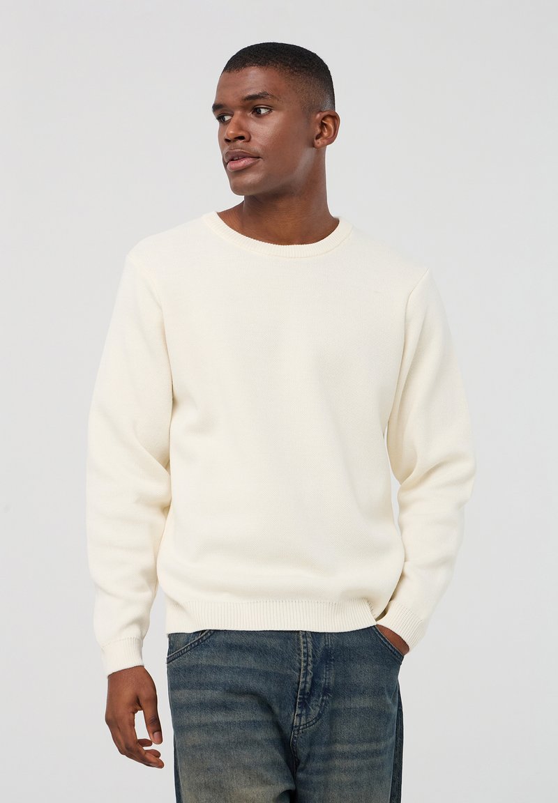 Maglione a maglia colore crema con colletto tondo, maniche lunghe e polsini a coste; indossato con jeans in denim blu; texture liscia, design semplice.